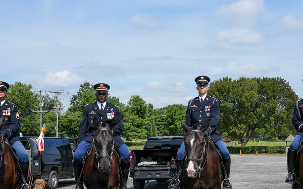 Four uniformed military or ceremonial personnel are mounted on dark bay horses in a gravel parking area. All four riders wear formal dress uniforms with medals, ribbons, and peaked caps, suggesting a ceremonial or parade context. The setting appears to be an outdoor staging area with vehicles, trees, and open sky in the background.