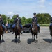 Four uniformed military or ceremonial personnel are mounted on dark bay horses in a gravel parking area. All four riders wear formal dress uniforms with medals, ribbons, and peaked caps, suggesting a ceremonial or parade context. The setting appears to be an outdoor staging area with vehicles, trees, and open sky in the background.