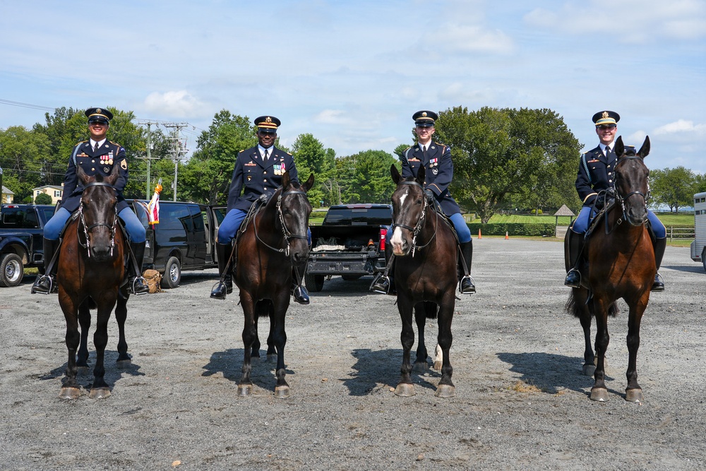 Four military personnel in dress uniforms are mounted on dark bay horses in a gravel parking area. The soldiers are wearing U.S. Army ceremonial uniforms with service ribbons and medals, suggesting this is a formal or ceremonial occasion. The setting appears to be an outdoor staging area with vehicles, trees, and open sky in the background.