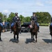 Four military personnel in dress uniforms are mounted on dark bay horses in a gravel parking area. The soldiers are wearing U.S. Army ceremonial uniforms with service ribbons and medals, suggesting this is a formal or ceremonial occasion. The setting appears to be an outdoor staging area with vehicles, trees, and open sky in the background.