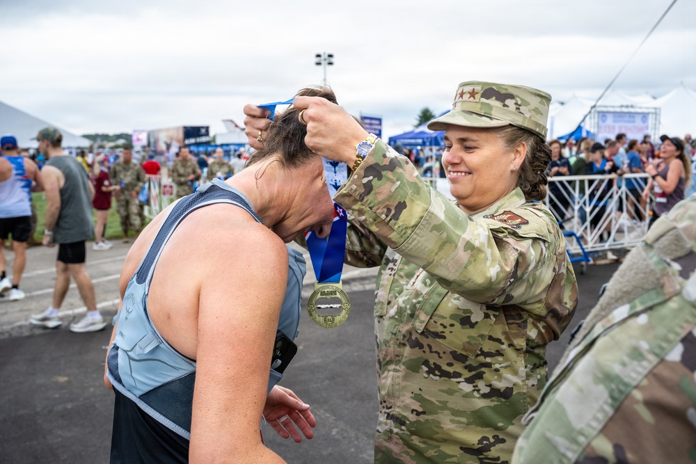 A military service member in OCP (Operational Camouflage Pattern) uniform is placing a finisher's medal around the neck of a race participant at what appears to be a military-sponsored running event. The soldier is smiling warmly as she lowers the medal over the runner's bowed head. The background shows a large outdoor event venue with tents, barriers, crowds, and other military personnel.