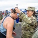 A military service member in OCP (Operational Camouflage Pattern) uniform is placing a finisher's medal around the neck of a race participant at what appears to be a military-sponsored running event. The soldier is smiling warmly as she lowers the medal over the runner's bowed head. The background shows a large outdoor event venue with tents, barriers, crowds, and other military personnel.