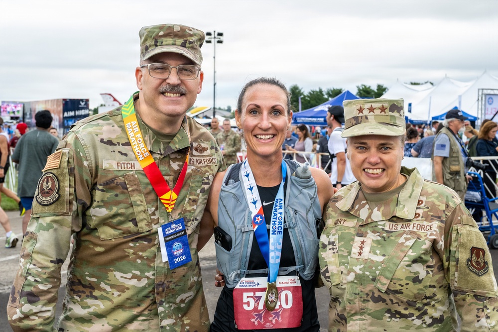 Three individuals pose together at the Air Force Marathon event, two of whom are wearing U.S. Air Force camouflage uniforms and one is a civilian runner wearing race bib number 5710 and a finisher's medal. The left uniformed individual has a name tag reading 'FITCH' and is wearing a marathon medal and 'Honored Guest 2026' lanyard, while the right uniformed individual has three stars on their cap indicating a Lieutenant General rank with a name tag partially reading 'ARRY'. The event appears to be taking place outdoors with vendor tents and crowds visible in the background.