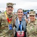 Three individuals pose together at the Air Force Marathon event, two of whom are wearing U.S. Air Force camouflage uniforms and one is a civilian runner wearing race bib number 5710 and a finisher's medal. The left uniformed individual has a name tag reading 'FITCH' and is wearing a marathon medal and 'Honored Guest 2026' lanyard, while the right uniformed individual has three stars on their cap indicating a Lieutenant General rank with a name tag partially reading 'ARRY'. The event appears to be taking place outdoors with vendor tents and crowds visible in the background.