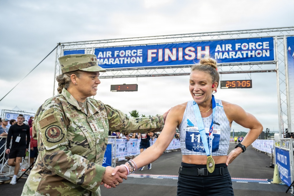 A U.S. Air Force officer in camouflage uniform congratulates a female marathon finisher at the Air Force Marathon finish line. The runner, wearing bib number 11 with the name 'Kayl', has just completed the race with a finish time of 2:57:05 visible on the clock. The runner is wearing a finisher's medal and both individuals are shaking hands and smiling.