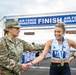 A U.S. Air Force officer in camouflage uniform congratulates a female marathon finisher at the Air Force Marathon finish line. The runner, wearing bib number 11 with the name 'Kayl', has just completed the race with a finish time of 2:57:05 visible on the clock. The runner is wearing a finisher's medal and both individuals are shaking hands and smiling.