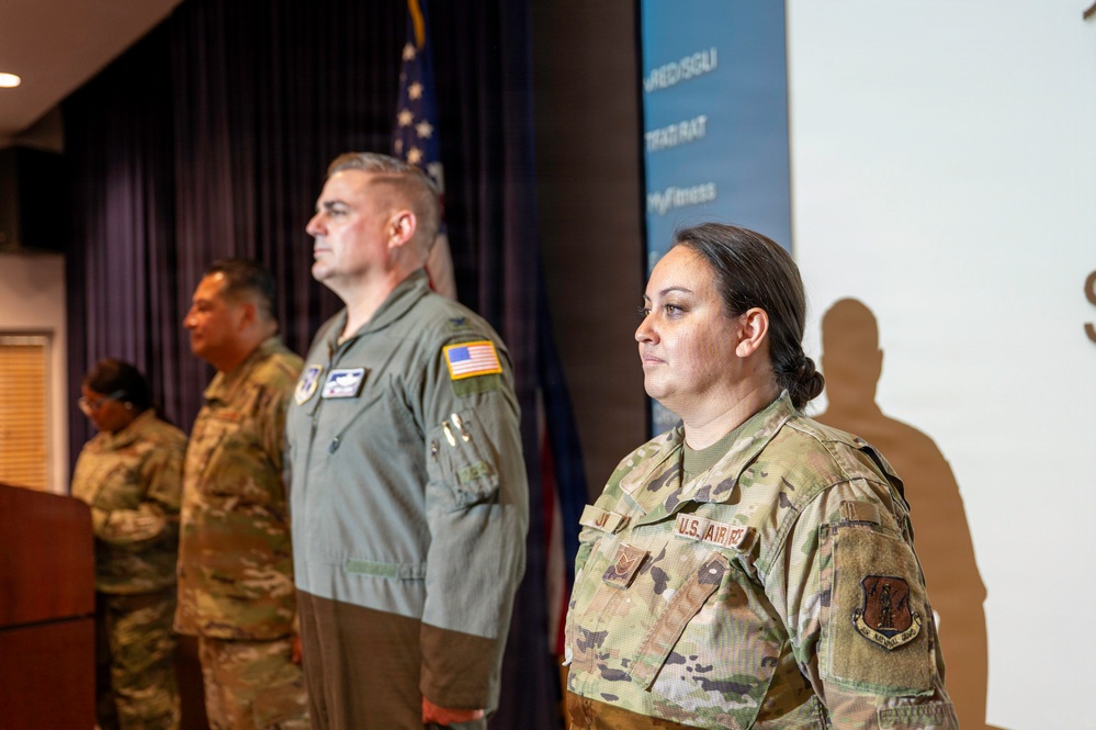 Several U.S. Air Force personnel are standing at attention in what appears to be an official ceremony or briefing room. The group includes individuals in both OCP (Operational Camouflage Pattern) uniforms and a flight suit, suggesting mixed roles. An American flag and a blue banner with partial text are visible in the background.