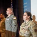 Several U.S. Air Force personnel are standing at attention in what appears to be an official ceremony or briefing room. The group includes individuals in both OCP (Operational Camouflage Pattern) uniforms and a flight suit, suggesting mixed roles. An American flag and a blue banner with partial text are visible in the background.
