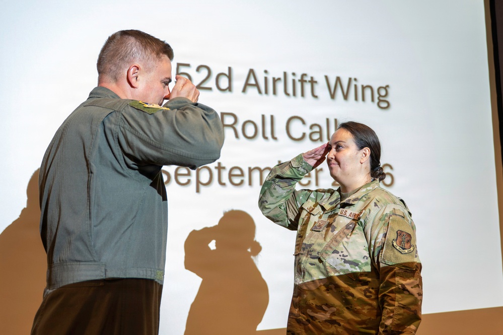 Two U.S. Air Force military personnel are exchanging salutes during a formal ceremony at the 52d Airlift Wing Roll Call event in September. The individual on the left is wearing an olive/flight suit uniform, while the individual on the right is in OCP (Operational Camouflage Pattern) uniform with visible rank insignia and a U.S. Air Force patch. Their shadows are projected dramatically on the presentation screen behind them.