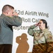 Two U.S. Air Force military personnel are exchanging salutes during a formal ceremony at the 52d Airlift Wing Roll Call event in September. The individual on the left is wearing an olive/flight suit uniform, while the individual on the right is in OCP (Operational Camouflage Pattern) uniform with visible rank insignia and a U.S. Air Force patch. Their shadows are projected dramatically on the presentation screen behind them.