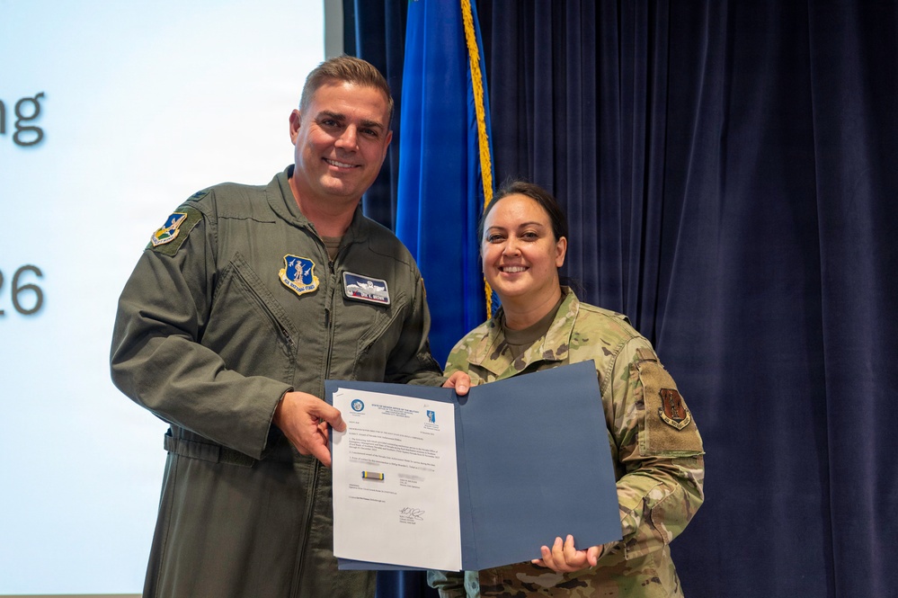 Two U.S. Air Force military personnel are posing together while holding an official certificate or award document in a blue folder. The individual on the left is wearing an olive green flight suit, while the individual on the right is wearing an OCP (Operational Camouflage Pattern) uniform. This appears to be a formal military recognition or award ceremony.