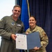 Two U.S. Air Force military personnel are posing together while holding an official certificate or award document in a blue folder. The individual on the left is wearing an olive green flight suit, while the individual on the right is wearing an OCP (Operational Camouflage Pattern) uniform. This appears to be a formal military recognition or award ceremony.