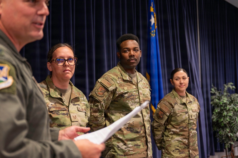 A U.S. Air Force ceremony is taking place in a formal indoor setting. A senior officer in a flight suit is reading from a document while three uniformed Airmen in OCP (Operational Camouflage Pattern) uniforms stand at attention. A blue Air Force flag with a star is visible in the background.
