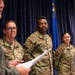 A U.S. Air Force ceremony is taking place in a formal indoor setting. A senior officer in a flight suit is reading from a document while three uniformed Airmen in OCP (Operational Camouflage Pattern) uniforms stand at attention. A blue Air Force flag with a star is visible in the background.