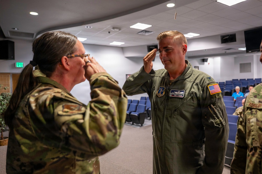 Two U.S. Air Force personnel are exchanging salutes in what appears to be an indoor auditorium or briefing room setting. The individual on the right is wearing a green flight suit with an American flag patch and military insignia, while the individual on the left is wearing an OCP (Operational Camouflage Pattern) uniform. This appears to be a formal military ceremony, possibly a promotion, change of command, or retirement event.