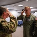 Two U.S. Air Force personnel are exchanging salutes in what appears to be an indoor auditorium or briefing room setting. The individual on the right is wearing a green flight suit with an American flag patch and military insignia, while the individual on the left is wearing an OCP (Operational Camouflage Pattern) uniform. This appears to be a formal military ceremony, possibly a promotion, change of command, or retirement event.