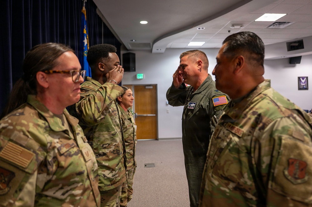 Military personnel in a formal indoor setting are exchanging salutes, suggesting a ceremony or official greeting. A mix of individuals wearing OCP (Operational Camouflage Pattern) uniforms and one person in a green flight suit are present. A blue flag, likely a U.S. Air Force or state flag, is visible in the background.