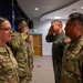 Military personnel in a formal indoor setting are exchanging salutes, suggesting a ceremony or official greeting. A mix of individuals wearing OCP (Operational Camouflage Pattern) uniforms and one person in a green flight suit are present. A blue flag, likely a U.S. Air Force or state flag, is visible in the background.