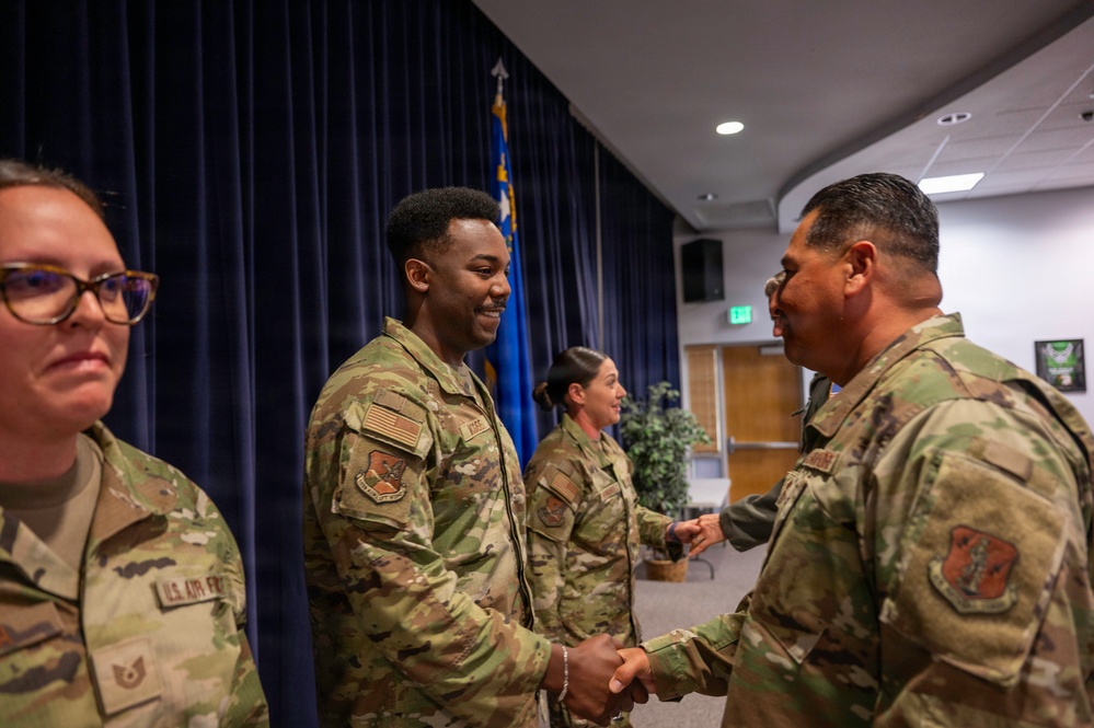 A formal military ceremony or recognition event is taking place in what appears to be a U.S. Air Force auditorium or meeting room. A senior military member is shaking hands with a younger Airman, suggesting a commendation, award, or greeting line. All personnel are wearing OCP (Operational Camouflage Pattern) uniforms with U.S. Air Force insignia.