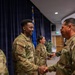 A formal military ceremony or recognition event is taking place in what appears to be a U.S. Air Force auditorium or meeting room. A senior military member is shaking hands with a younger Airman, suggesting a commendation, award, or greeting line. All personnel are wearing OCP (Operational Camouflage Pattern) uniforms with U.S. Air Force insignia.