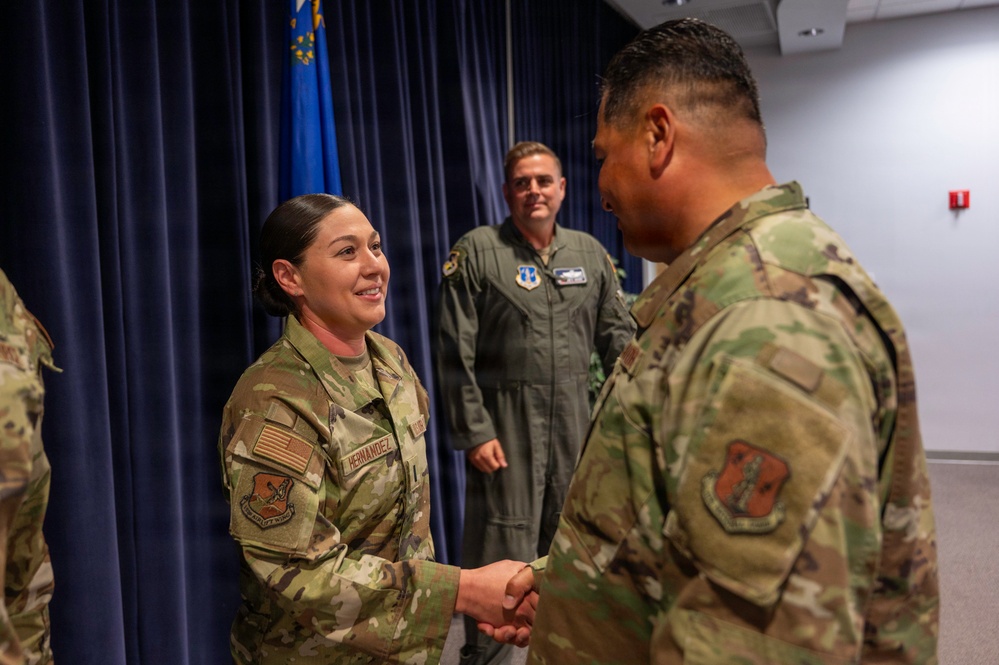 A U.S. Air Force ceremony showing a female airman named Hernandez shaking hands with another service member in OCP (Operational Camouflage Pattern) uniforms. A third individual in an olive green flight suit stands in the background observing the handshake. The setting appears to be an indoor military ceremony room with a blue curtain and U.S. Air Force flag visible.