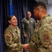 A U.S. Air Force ceremony showing a female airman named Hernandez shaking hands with another service member in OCP (Operational Camouflage Pattern) uniforms. A third individual in an olive green flight suit stands in the background observing the handshake. The setting appears to be an indoor military ceremony room with a blue curtain and U.S. Air Force flag visible.