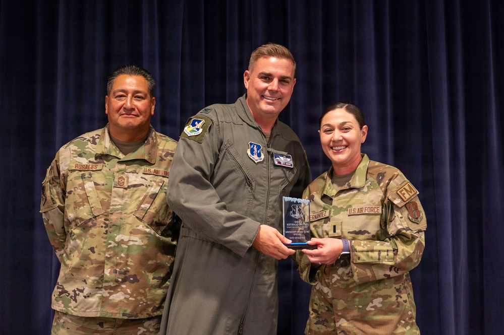 Three U.S. Air Force personnel are posing for a formal photo during what appears to be an awards ceremony. The central figure in a flight suit and the person on the right are jointly holding a glass/crystal award plaque. The individuals are identified by name tapes as Rosales (left) and Hernandez (right).