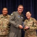Three U.S. Air Force personnel are posing for a formal photo during what appears to be an awards ceremony. The central figure in a flight suit and the person on the right are jointly holding a glass/crystal award plaque. The individuals are identified by name tapes as Rosales (left) and Hernandez (right).