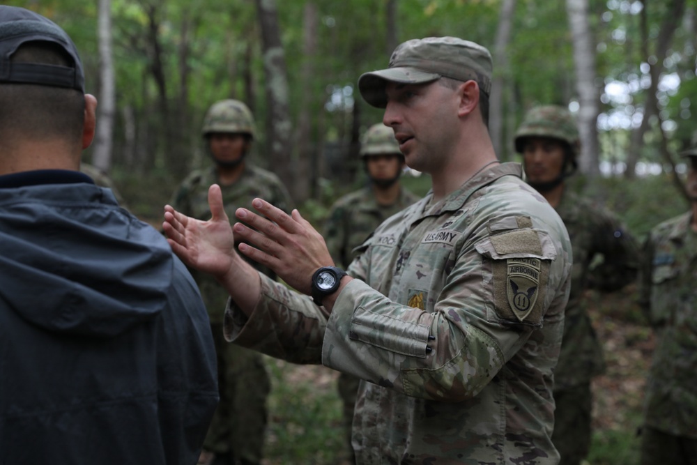 A U.S. Army soldier wearing OCP (Operational Camouflage Pattern) uniform with Airborne and 11th patch insignia is briefing or instructing a group in a wooded outdoor setting. Several soldiers in different camouflage uniforms, appearing to be from a foreign military, are listening in the background. The U.S. soldier is gesturing with both hands, suggesting active instruction or explanation.