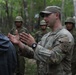 A U.S. Army soldier wearing OCP (Operational Camouflage Pattern) uniform with Airborne and 11th patch insignia is briefing or instructing a group in a wooded outdoor setting. Several soldiers in different camouflage uniforms, appearing to be from a foreign military, are listening in the background. The U.S. soldier is gesturing with both hands, suggesting active instruction or explanation.