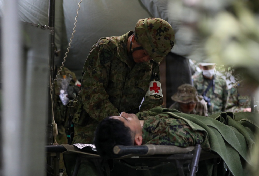 A military medical exercise or field treatment scenario showing a medic attending to a casualty on a stretcher. The standing soldier wears a Red Cross armband, identifying them as a combat medic, and is leaning over a patient in camouflage uniform lying on a field stretcher. Additional personnel in similar camouflage uniforms are visible in the background near what appears to be a military tent or vehicle.