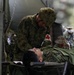 A military medical exercise or field treatment scenario showing a medic attending to a casualty on a stretcher. The standing soldier wears a Red Cross armband, identifying them as a combat medic, and is leaning over a patient in camouflage uniform lying on a field stretcher. Additional personnel in similar camouflage uniforms are visible in the background near what appears to be a military tent or vehicle.