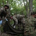 Military personnel from what appear to be two different armed forces are conducting a joint medical training exercise in a wooded area. Soldiers are working together to deploy or handle a litter/stretcher, suggesting a casualty evacuation drill. The presence of medical armbands and latex gloves indicates this is a combat medical training scenario.
