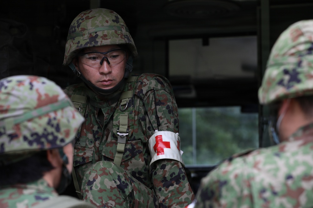 A military medic wearing a Red Cross armband is visible among a group of soldiers in camouflage uniforms and helmets. The central figure wears protective eyewear and appears to be engaged in a briefing or medical exercise. The camouflage pattern and equipment are consistent with Japan Ground Self-Defense Force (JGSDF) uniforms.