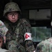 A military medic wearing a Red Cross armband is visible among a group of soldiers in camouflage uniforms and helmets. The central figure wears protective eyewear and appears to be engaged in a briefing or medical exercise. The camouflage pattern and equipment are consistent with Japan Ground Self-Defense Force (JGSDF) uniforms.