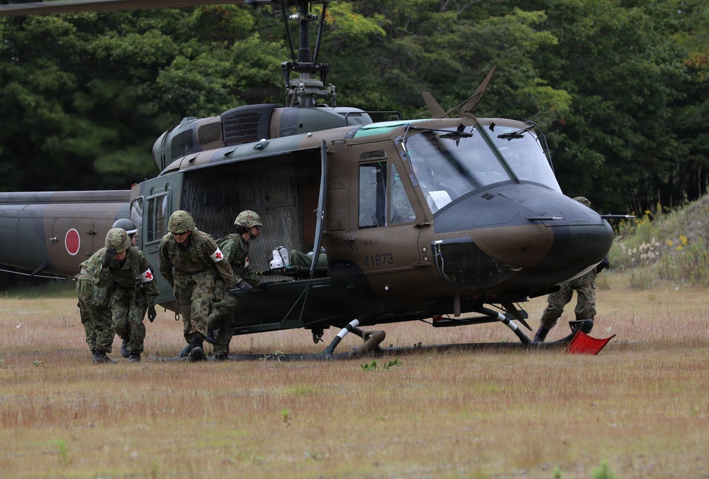 A military medical evacuation exercise is taking place on a grassy field. Soldiers wearing camouflage uniforms with Red Cross insignia are assisting a casualty near a Japanese Ground Self-Defense Force UH-1 Iroquois helicopter (tail number 41873). A second helicopter with a Japanese rising sun roundel is visible in the background.