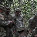 A group of U.S. Army soldiers are gathered under a camouflage netting in a wooded outdoor environment. Several soldiers appear to be reviewing documents or a clipboard while a senior NCO observes. The setting suggests a field training exercise or tactical operations briefing.