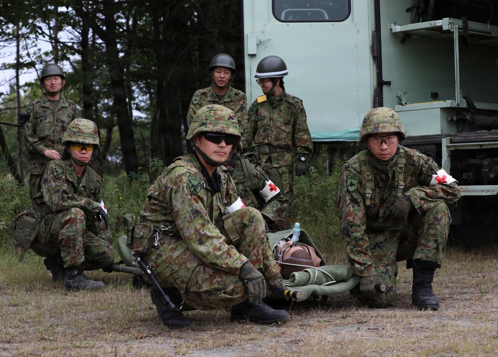 A group of military personnel in camouflage uniforms are conducting what appears to be a medical training exercise outdoors. Two soldiers wearing red cross armbands are attending to a simulated casualty lying on a stretcher on the ground. The scene takes place near a military vehicle with trees visible in the background.