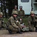 A group of military personnel in camouflage uniforms are conducting what appears to be a medical training exercise outdoors. Two soldiers wearing red cross armbands are attending to a simulated casualty lying on a stretcher on the ground. The scene takes place near a military vehicle with trees visible in the background.