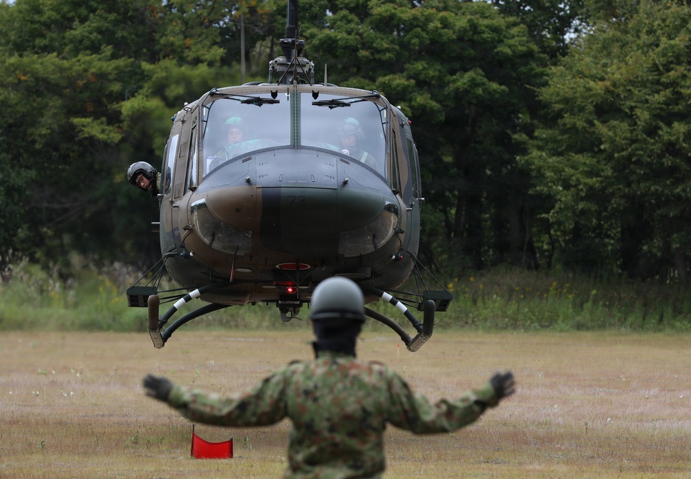 A military helicopter, appearing to be a UH-1 Iroquois (Huey) variant in olive drab livery, is hovering low over a grassy field during what appears to be a landing or takeoff operation. A ground crew member in camouflage uniform and helmet is directing the aircraft with outstretched arms. The setting appears to be a military training or operational area surrounded by dense trees.