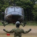 A military helicopter, appearing to be a UH-1 Iroquois (Huey) variant in olive drab livery, is hovering low over a grassy field during what appears to be a landing or takeoff operation. A ground crew member in camouflage uniform and helmet is directing the aircraft with outstretched arms. The setting appears to be a military training or operational area surrounded by dense trees.