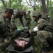 Military personnel in camouflage uniforms are conducting a combat casualty care training exercise in a forested area. A simulated casualty (shirtless individual) is lying on a stretcher while multiple soldiers attend to him, with at least one wearing blue medical gloves. One soldier displays a red cross armband, indicating a medic role, suggesting this is a joint military medical training exercise.