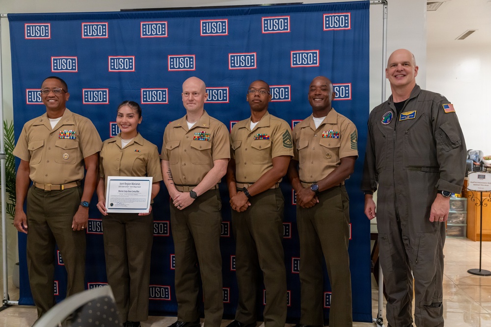 Six military personnel pose for a group photo in front of a USO-branded backdrop. One individual is holding a framed certificate or award, suggesting this is a recognition or award ceremony. The group includes members in U.S. Marine Corps service uniforms and one individual in an olive/gray flight suit.