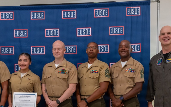 Six military personnel pose for a group photo in front of a USO-branded backdrop. One individual is holding a framed certificate or award, suggesting this is a recognition or award ceremony. The group includes members in U.S. Marine Corps service uniforms and one individual in an olive/gray flight suit.