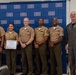 Six military personnel pose for a group photo in front of a USO-branded backdrop. One individual is holding a framed certificate or award, suggesting this is a recognition or award ceremony. The group includes members in U.S. Marine Corps service uniforms and one individual in an olive/gray flight suit.