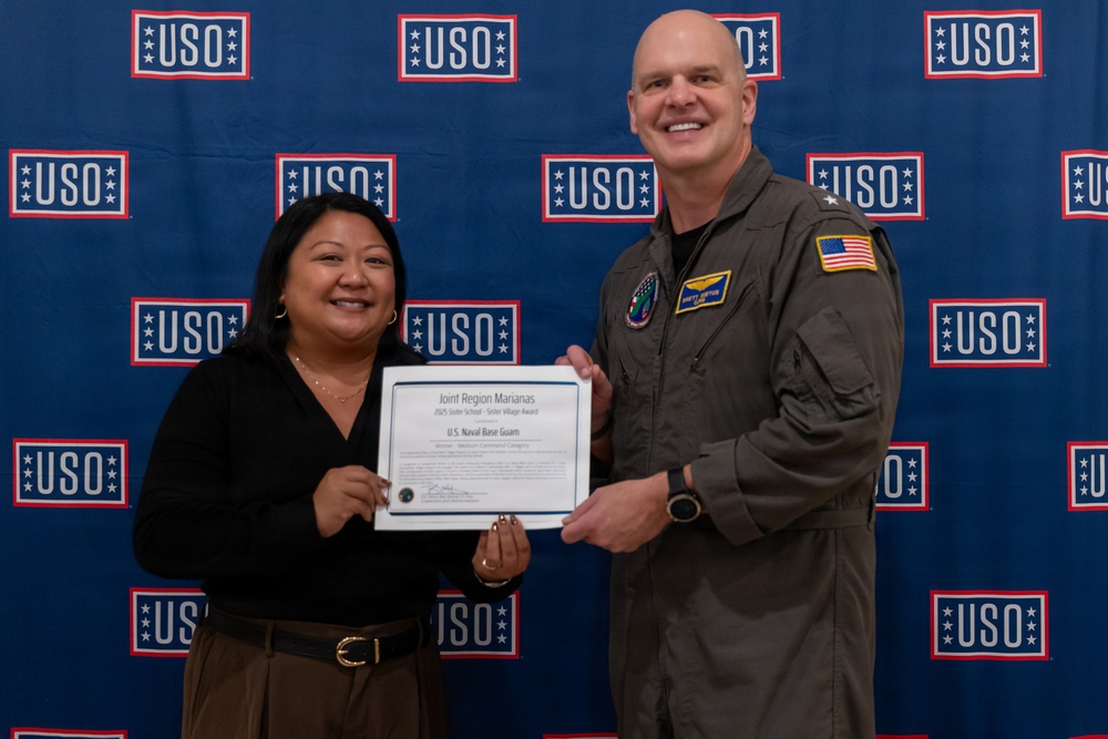 Two individuals are posing together in front of a USO-branded backdrop while holding a certificate. The certificate reads 'Joint Region Marianas' and references 'U.S. Naval Base Guam,' suggesting an award presentation ceremony. The military officer is wearing an olive green flight suit with an American flag patch, while the civilian woman is dressed in business casual attire.
