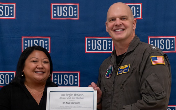 Two individuals are posing together in front of a USO-branded backdrop while holding a certificate. The certificate reads 'Joint Region Marianas' and references 'U.S. Naval Base Guam,' suggesting an award presentation ceremony. The military officer is wearing an olive green flight suit with an American flag patch, while the civilian woman is dressed in business casual attire.