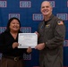 Two individuals are posing together in front of a USO-branded backdrop while holding a certificate. The certificate reads 'Joint Region Marianas' and references 'U.S. Naval Base Guam,' suggesting an award presentation ceremony. The military officer is wearing an olive green flight suit with an American flag patch, while the civilian woman is dressed in business casual attire.