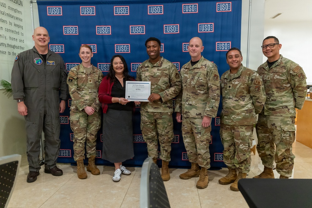 A group of seven individuals pose in front of a USO backdrop, with two people in the center holding a certificate or award document. Six of the individuals are wearing military camouflage (OCP) uniforms or a flight suit, while one civilian woman in a red jacket and black skirt stands at the center. This appears to be a formal recognition or award ceremony at a USO location.