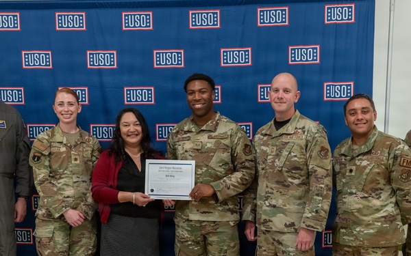 A group of seven individuals pose in front of a USO backdrop, with two people in the center holding a certificate or award document. Six of the individuals are wearing military camouflage (OCP) uniforms or a flight suit, while one civilian woman in a red jacket and black skirt stands at the center. This appears to be a formal recognition or award ceremony at a USO location.